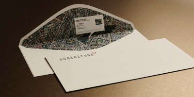 Geöffneter weißer Briefumschlag der Marke Augenzeuge: Die Innenseite ist vollflächig mit einem detaillierten Stadtplan-Ausschnitt (Google Maps Stil) bedruckt, der als Wegbeschreibung dient.