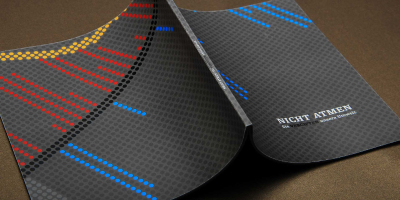 Ein dunkelgraues, quadratisches Magazin liegt weit aufgeschlagen auf einer braunen Fläche. Das Cover zeigt ein minimalistisches Punktraster mit blau-roten Farbakzenten und den Titel ‚NICHT ATMEN – Sie vergiften unsere Umwelt‘.