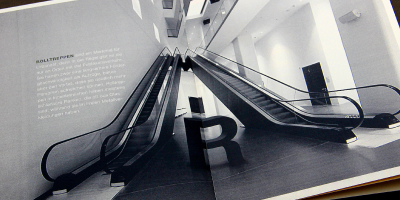Dynamische Doppelseite zum Thema Urbanität. Eine Weitwinkel-Fotografie blickt von unten auf zwei parallel verlaufende Rolltreppen in einem modernen, lichtdurchfluteten Gebäude. Ein großes schwarzes ‚R‘ ist zentral im unteren Bildbereich platziert.