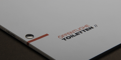 Close-up des Magazin-Covers mit dem Titel „Öffentliche Toiletten //“. Minimalistisches Design auf lichtgrauem Grund mit serifenloser Typografie. Eine metallische Buchschraube ist als Teil der Bindung sichtbar.