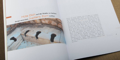 Magazin-Innenseite über die Geschichte antiker Toiletten. Links ein Foto historischer Steinlatrinen, darüber die Headline „Hüte Dich...“ in Orange und Schwarz. Rechts ein strukturierter Textblock in einer modernen Sans-Serif. Das Layout besticht durch viel Weißraum und eine klare Hierarchie.