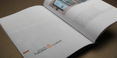 Geöffnete Doppelseite mit dem Titel „Zwischen Fäkalien & Luxus“. Ein asymmetrisches Raster ordnet das Foto einer modernen Anlage oben rechts und die Typografie unten links an. Ein oranges Ampersand dient als zentrales gestalterisches Verbindungselement.