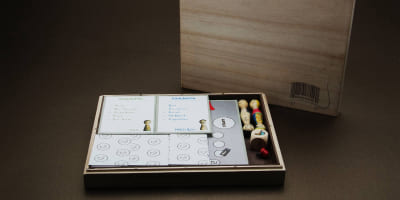 Holzkiste mit Moosgummi-Einlage, darin Spielfiguren, Karten, Würfel und Spielbrett der Einkaufsrallye; die Kiste schließt mit Magneten.