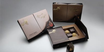 Mehrere hochwertige Pralinenverpackungen in drei Sorten — Whisky‑Sahne, Cognac‑Sahne und Marc de Champagne — sind aufgeklappt zu sehen. Jede Box zeigt acht Pralinen, davon vier in einem kleinen tragbaren Karton.