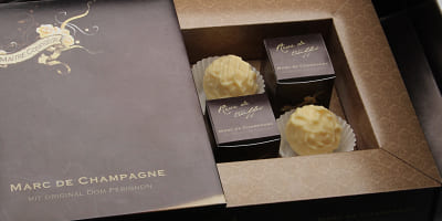 Eine Nahaufnahme einer edlen Pralinenverpackung mit weißen Marc‑de‑Champagne‑Trüffeln. Die Pralinen liegen in Papierkapseln, umgeben von einer dunkelbraunen, goldverzierten Box; im Hintergrund sind kleine Einzelkartons sichtbar.