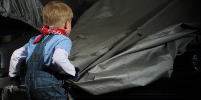 Ein kleiner Junge mit blonden Haaren, gekleidet in eine Latzhose und ein rotes Halstuch, zieht in einer dunklen Garage eine graue Schutzplane von einem Auto. Er steht mit dem Rücken zur Kamera und scheint gespannt auf das zu sein, was sich darunter verbirgt.