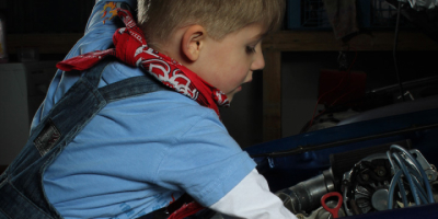 Der Junge beugt sich tief in den Motorraum des blauen Wagens. Seine Hände sind bei der Arbeit am Motor im Schatten verschwunden. Sein Gesichtsausdruck ist ernst und voller Fokus, während er die Rolle eines Mechanikers einnimmt.