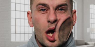 Eine Person in einem Innenraum, deren Gesicht seitlich durch kräftige Luftkraft eingedrückt und verzogen wirkt.