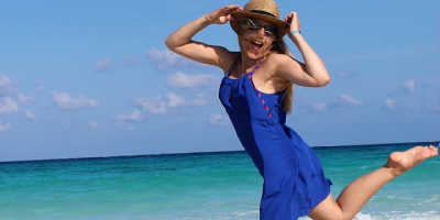 Eine Person springt am Strand in die Luft, trägt ein blaues Kleid und einen Strohhut. Hinter ihr sind Meer und blauer Himmel zu sehen.