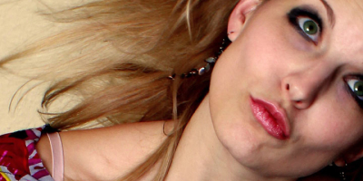 Eine Person mit langen blonden Haaren trägt ein farbenfrohes Oberteil. Die Haare wehen zur Seite, das Gesicht zeigt einen ausdrucksstarken, dynamischen Blick.