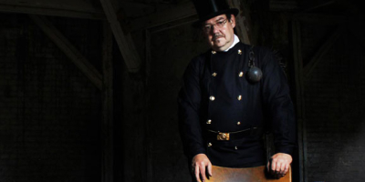 Eine Person in traditioneller Schornsteinfeger‑Kleidung steht in einem Dachboden. Sie trägt einen schwarzen Mantel, einen Zylinder und hat Ruß im Gesicht. Ein Schornsteinfegerwerkzeug hängt über der Schulter.